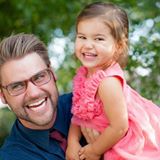 A smiling man holding a joyful young girl outdoors.
