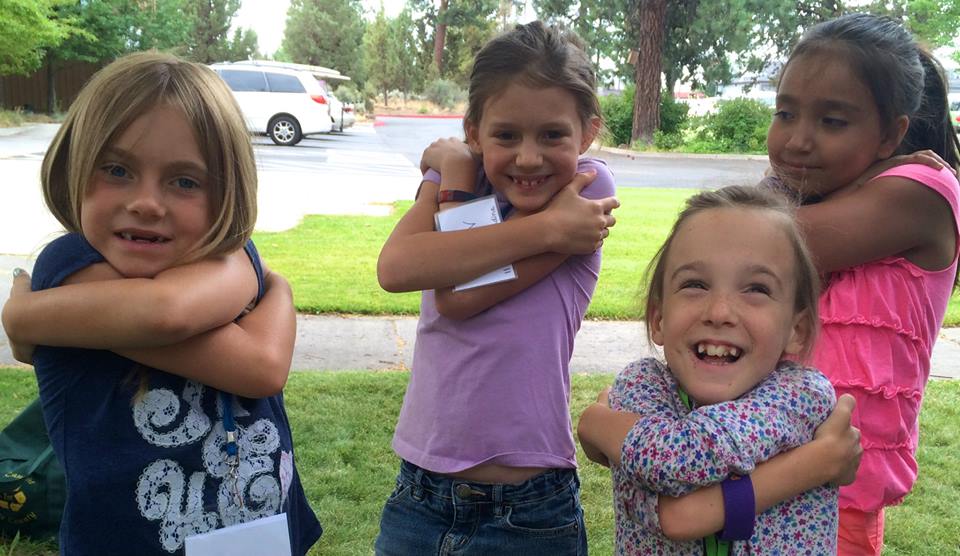 Four young girls stand outside, smiling and hugging themselves. The setting is a grassy area with some trees and cars in the background.