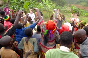 A large group of people dancing outdoors, with their arms raised, in a celebratory atmosphere. The background consists of green banana plants and a rural landscape.