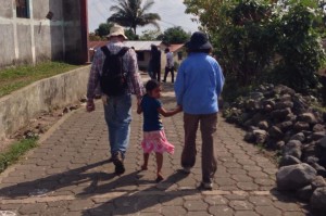 A child walking hand-in-hand with two adults on a paved path in a rural setting.