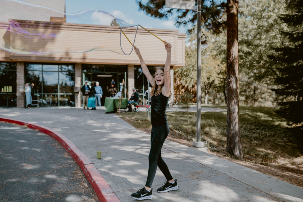A girl in a black outfit joyfully swings a bubble wand above her head outside a church event.