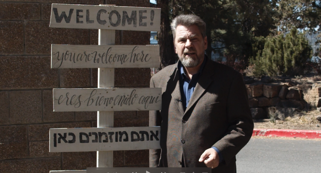 A man with short, graying hair and a beard stands in front of a wooden welcome sign with multiple languages, smiling at the camera.