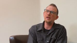 A man with short, graying hair and a beard sits on a couch, looking slightly upwards, wearing glasses and a checkered shirt.