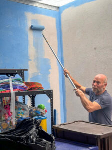 A man with short, graying hair and a beard is painting a wall with a blue roller. He is standing beside a shelf filled with toys and materials.