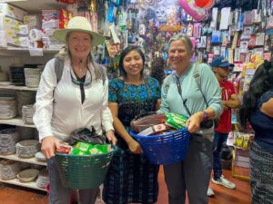 Three women stand in a shop filled with various items. Two women are holding baskets filled with supplies, while a third woman stands between them. Brightly colored packages and items are visible on the shelves behind them.