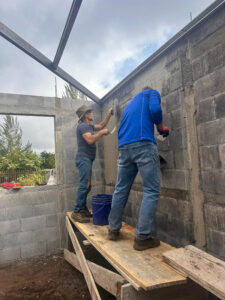 Two men working on a wall, one applying cement and the other mixing it, with a foundation visible in the background.