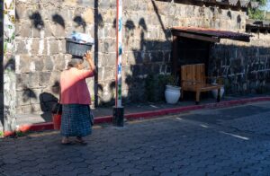 An elderly woman wearing a bright pink top and a traditional patterned skirt walks along a stone wall, balancing a black basket of items on her head.