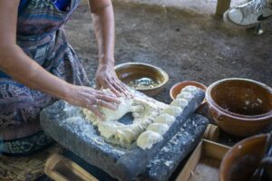 A woman working with dough on a flat stone surface, with bowls nearby, in a rustic setting.