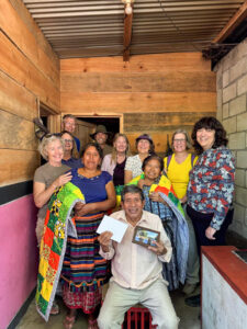 A group of people poses indoors, holding colorful blankets and items. A man in the front displays a photo and a letter.