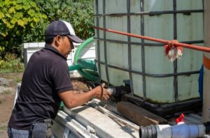 A man wearing a black polo shirt and a baseball cap is operating equipment next to a large container on the back of a truck.